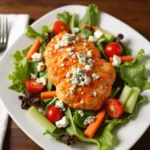 A Buffalo chicken salad with crispy chicken tenders drizzled in Buffalo sauce, topped with blue cheese crumbles, served on a bed of fresh greens, cherry tomatoes, celery, and carrots.