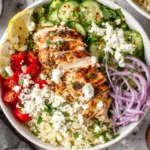 Greek Chicken Bowls with grilled chicken, rice, cucumber, cherry tomatoes, red onion, feta cheese, and lemon slices in a healthy Mediterranean meal