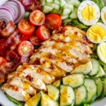 Grilled Chicken Cobb Salad with Honey Dijon featuring sliced grilled chicken, crispy bacon, cherry tomatoes, eggs, and fresh veggies in a white bowl.