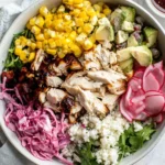 Colorful rotisserie chicken salad bowl with corn, avocado, pickled radish, and shredded chicken on a bed of greens.