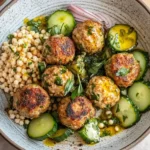 Herby Greek Chicken Meatballs served with giant couscous, cucumber salad, and fresh herbs in a rustic bowl