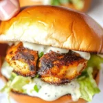 Hand holding an Alabama White Blackened Chicken Slider with crispy blackened chicken, creamy white BBQ sauce, and lettuce on a toasted bun.
