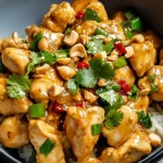 Close-up of Thai Peanut Chicken Stir-Fry with chopped peanuts, fresh cilantro, and chili over white rice in a dark bowl.