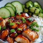 Sticky Honey Gochujang Chicken served with rice and cucumbers in a bowl, topped with green onions and sesame seeds.