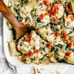 Spinach Artichoke Chicken Bake in a white baking dish with creamy sauce, artichoke hearts, spinach, and red peppers