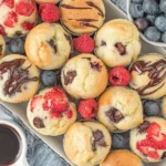 Freshly baked Pancake Mini Muffins with chocolate drizzle, blueberries, raspberries, and strawberries, served with syrup for dipping.