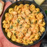 Creamy CrockPot Chicken Tortellini in a black bowl garnished with fresh herbs and surrounded by shredded cheese and basil leaves.