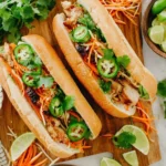 Grilled chicken Banh Mi Sandwich topped with pickled carrots, fresh cilantro, jalapeños, and lime wedges on a wooden board.