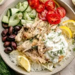 Slow Cooker Greek Chicken bowl with shredded chicken, rice, olives, cucumbers, tomatoes, lemon, and tzatziki sauce.
