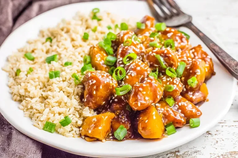 Slow Cooker Orange Chicken served with fluffy brown rice, coated in a glossy sweet-tangy orange sauce and topped with sesame seeds and sliced green onions on a white plate.