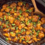 Slow Cooker Orange Chicken cooking in a crockpot, coated in a sticky orange glaze and topped with sesame seeds and scallions.