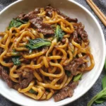 Bowl of Simple Black Pepper Beef Udon with tender beef slices, chewy noodles, and greens on a dark napkin background.