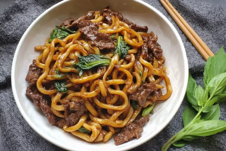 Bowl of Simple Black Pepper Beef Udon with tender beef slices, chewy noodles, and greens on a dark napkin background.