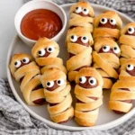 Plate of Mummy Pigs in a Blanket with candy eyes served with ketchup dipping sauce, a fun Halloween party snack.