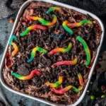 Colorful gummy worms on top of a rich chocolate pudding dirt cake layered with crushed Oreos and creamy frosting in a white baking dish.
