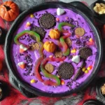 Halloween Dunkaroo Dip in a black cauldron topped with Oreos, gummy worms, candy pumpkins, and sprinkles surrounded by mini pumpkins and cauldrons of nuts.