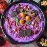 Colorful Halloween Dunkaroo Dip topped with Oreos, gummy worms, candy pumpkins, and festive sprinkles in a black cauldron bowl.