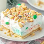 Slice of No Bake Christmas Cookie Lush topped with whipped cream, crushed cookies, and colorful holiday sprinkles on a white plate.