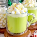 Glass mug of creamy green Grinch Hot Chocolate topped with whipped cream and mini marshmallows on a festive holiday table.