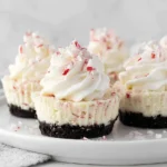 Peppermint Cheesecakes topped with whipped cream and crushed candy canes on a white plate, featuring a chocolate Oreo crust and creamy peppermint filling