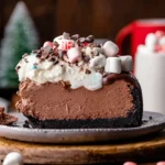 Slice of Hot Chocolate Cheesecake topped with whipped cream, mini marshmallows, chocolate shavings, and festive sprinkles on a plate.