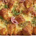 Croissant Bake in a white baking dish with golden croissants, melted Swiss and mozzarella, fresh herbs, and ribbons of ham.