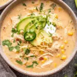 Slow Cooker Creamy Green Chile Chicken Enchilada Soup topped with avocado slices, shredded cheese, cilantro, and a jalapeño in a cozy bowl.