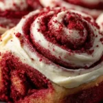 Close-up of freshly baked Red Velvet Cinnamon Rolls topped with creamy frosting and red velvet crumbs, showing their soft, swirled texture.