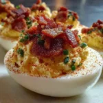 Million Dollar Bacon Deviled Eggs topped with crispy bacon, smoked paprika, and chives on a white serving plate.