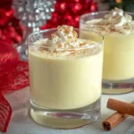 Homemade Eggnog in glass cups topped with whipped cream and cinnamon surrounded by holiday decorations.