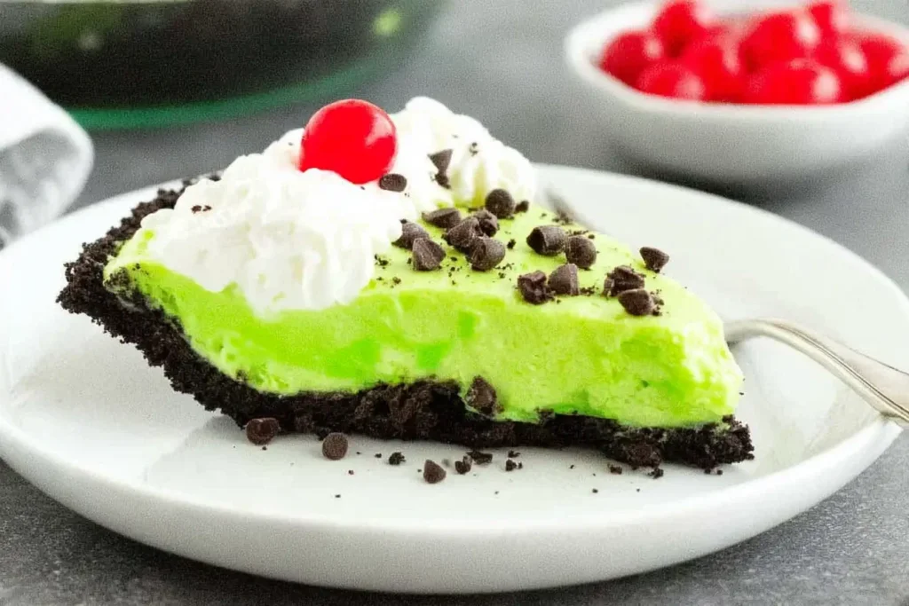 Slice of No Bake Grinch Pie with whipped cream, chocolate chips, and a cherry on an Oreo crust