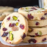 : Cranberry Pistachio Shortbread cookies stacked on parchment, showing buttery slices studded with dried cranberries and chopped pistachios.