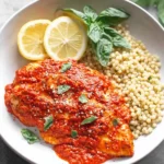 Mediterranean Chicken with Roasted Red Pepper Sauce served on a plate with pearl couscous, lemon slices, and fresh basil.