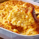 Golden-brown Corn Pudding in a white baking dish with a spoon lifting a creamy, custardy scoop filled with sweet corn kernels.