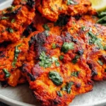 Paleo Tandoori Chicken thighs piled on a plate with charred edges, vibrant tandoori spices, and cilantro, served with a lemon wedge.