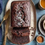 Chocolate Espresso Banana Bread loaf topped with chocolate chips and powdered sugar, sliced on a plate with coffee and bananas.