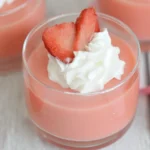 Strawberry Greek Yogurt Jello in a glass topped with whipped cream and fresh strawberry slices on a light wooden table.