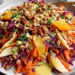 Crunchy Apple Carrot Salad with Creamy Citrus Dressing topped with pecans, orange slices, apples, shredded carrots, and red cabbage on a white plate.