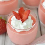 Strawberry Greek Yogurt Jello cups topped with whipped cream and sliced strawberries, arranged on a wooden surface.