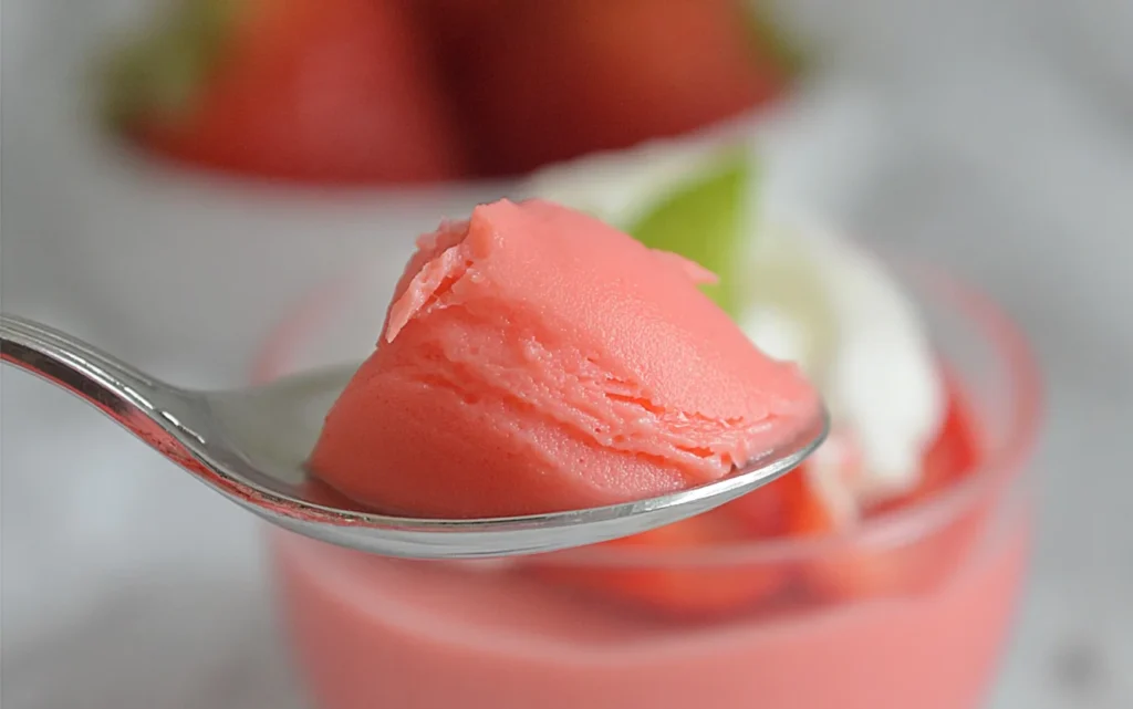 Spoonful of Strawberry Greek Yogurt Jello showing creamy, smooth texture with a blurred dessert cup and strawberry in the background.