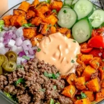 Sweet Potato Burger Bowl topped with creamy special sauce, featuring roasted sweet potatoes, seasoned ground beef, cucumbers, tomatoes, jalapeños, and red onion on lettuce.