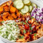 BBQ Chicken Bowls with roasted sweet potatoes, creamy coleslaw, tangy pickles, and saucy shredded chicken topped with fresh herbs.