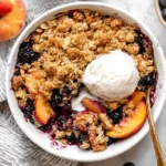 Blueberry Peach Crumble in a white baking dish with golden oat topping and a scoop of vanilla ice cream, surrounded by fresh peaches and blueberries
