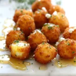 Crispy Goat Cheese Poppers with Honey on a white platter, golden fried bites drizzled with honey and sprinkled with herbs.