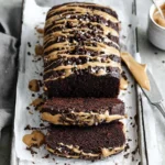 Chocolate Peanut Butter Banana Bread loaf sliced on parchment paper with creamy peanut butter drizzle and chocolate chips.
