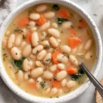 Bowl of Greek White Bean Soup With Garlic & Lemon with tender cannellini beans, carrots, herbs, and a lemon wedge on the side.