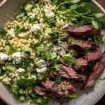 Grilled Skirt Steak with Lemon Herb Couscous Salad in a bowl with cucumber, fresh herbs, feta, and sliced grilled skirt steak.