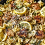 Lemon Chicken Pasta in a skillet with bowtie pasta, seared chicken, zucchini, parmesan, and fresh lemon slices.