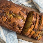 Chocolate Chip Banana Bread loaf sliced on a wooden board with melty chocolate chips and a cozy kitchen towel.