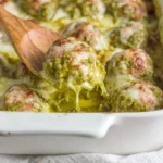 Close-up of Pesto Baked Turkey Meatballs on a wooden spoon, covered in basil pesto with stretchy melted mozzarella in a baking dish.